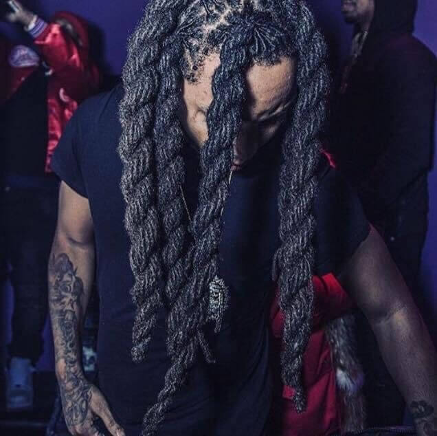 A man with Chunky Dreadlocks standing in front of a purple wall and tattoo in his hand